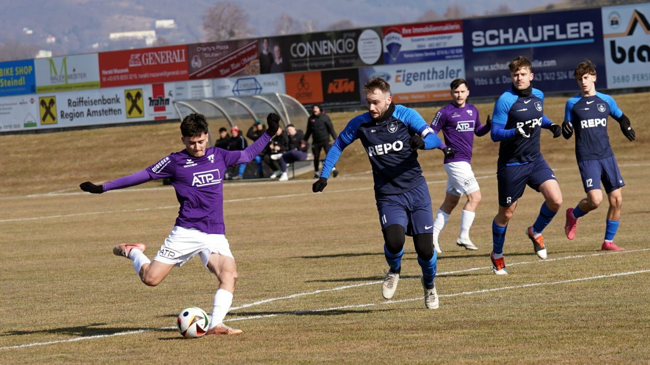 Ein Fußballspiel mit Spielern in lila und blauen Trikots, ein Spieler in Blau läuft auf den Ball zu, während der Spieler in Lila versucht, ihn zu blockieren.