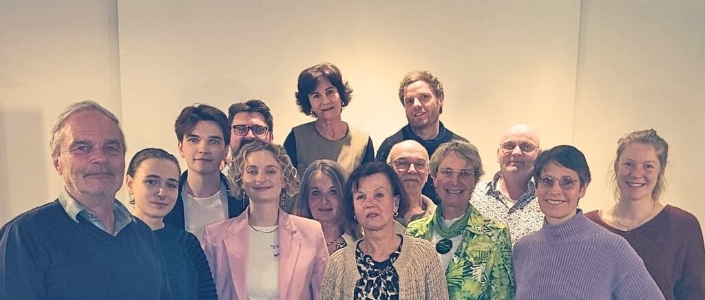 Eine Gruppe von Erwachsenen und jungen Menschen posiert für ein Foto. Die Gruppe besteht aus Männern und Frauen, die alle Freizeitkleidung tragen. Die Frau in der Mitte trägt eine Halskette und einen Blazer. Der Raum hat weiße Wände.