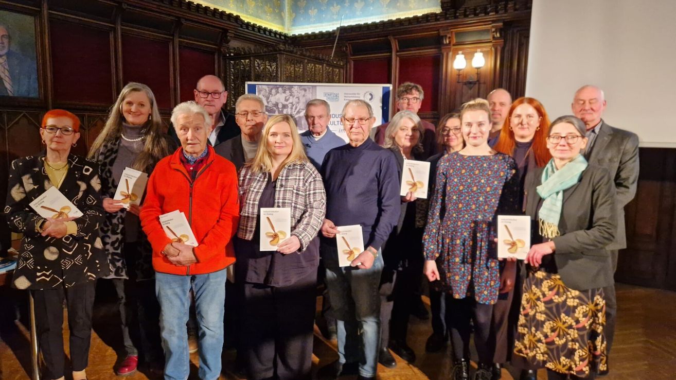Eine Gruppe von Menschen steht zusammen, hält Bücher und lächelt, in einem Raum mit Holzwänden und einem Banner im Hintergrund.