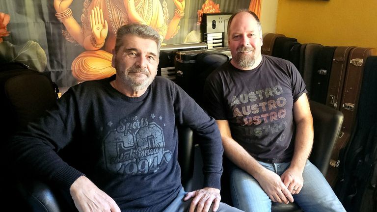 Zwei Männer sitzen zusammen in einem Studio. Einer trägt ein schwarzes T-Shirt mit 'The Sports' und Sternen, der andere ein schwarzes T-Shirt mit 'AUSTRO'. Dahinter ist eine große goldene Figur eines Gottes mit erhobenen Händen zu sehen.