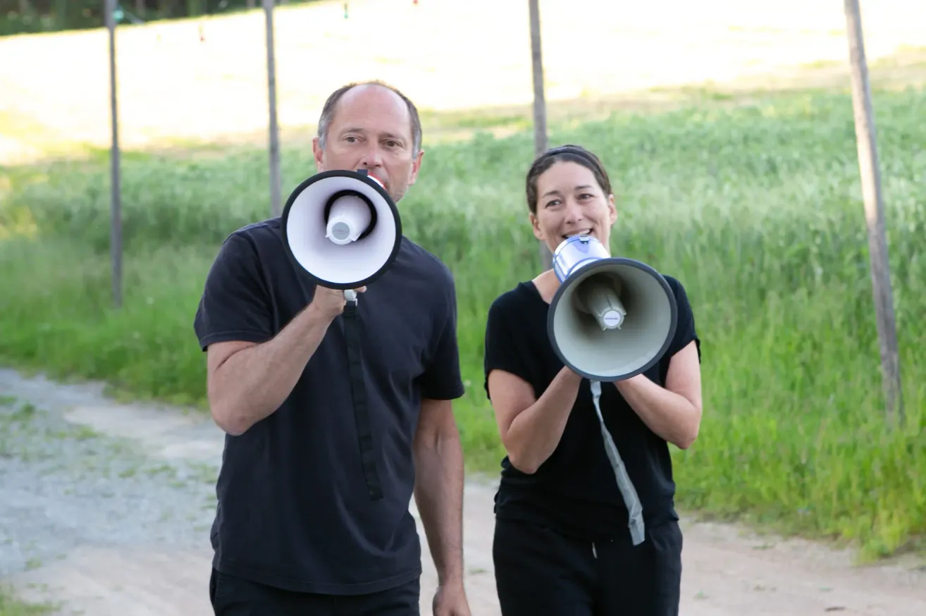 Ein Mann und eine Frau halten Megaphone und lächeln, stehen im Freien mit hohem Gras dahinter.