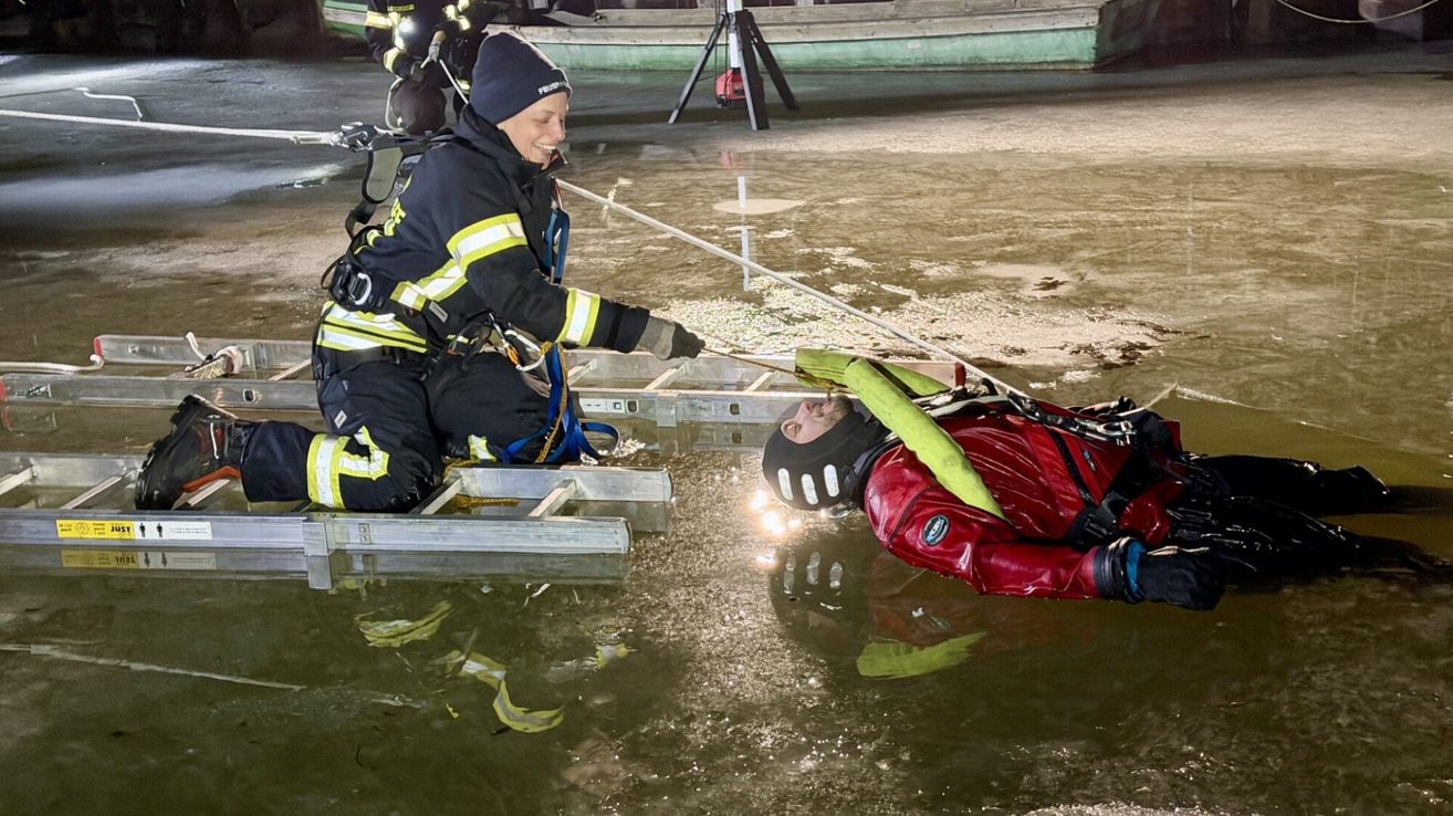 Ein Feuerwehrmann rettet eine Person in einem roten Anzug aus einem Gewässer mithilfe eines Seils. Beide tragen Helme und Sicherheitsausrüstung. Ein anderer Feuerwehrmann steht in der Nähe.