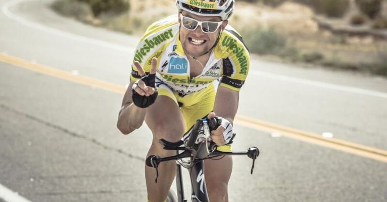 Ein Radfahrer in einem gelben Trikot mit dem Wort 'Wiesbauer' fährt auf einer Straße und lächelt, während er ein Friedenszeichen macht.