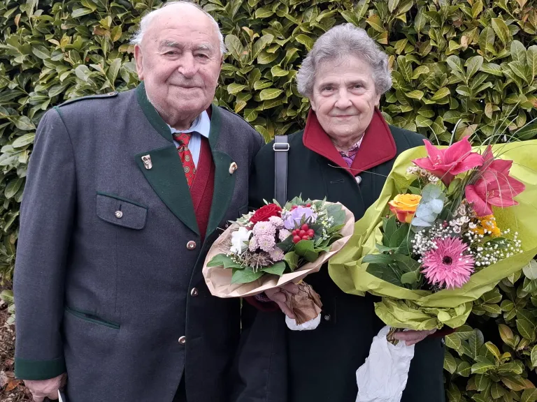 Ein älteres Paar steht lächelnd für ein Foto zusammen und hält Blumensträuße in den Händen. Der Mann trägt eine rote Krawatte und die Frau hat einen roten Kragen. Sie stehen vor einem Busch.