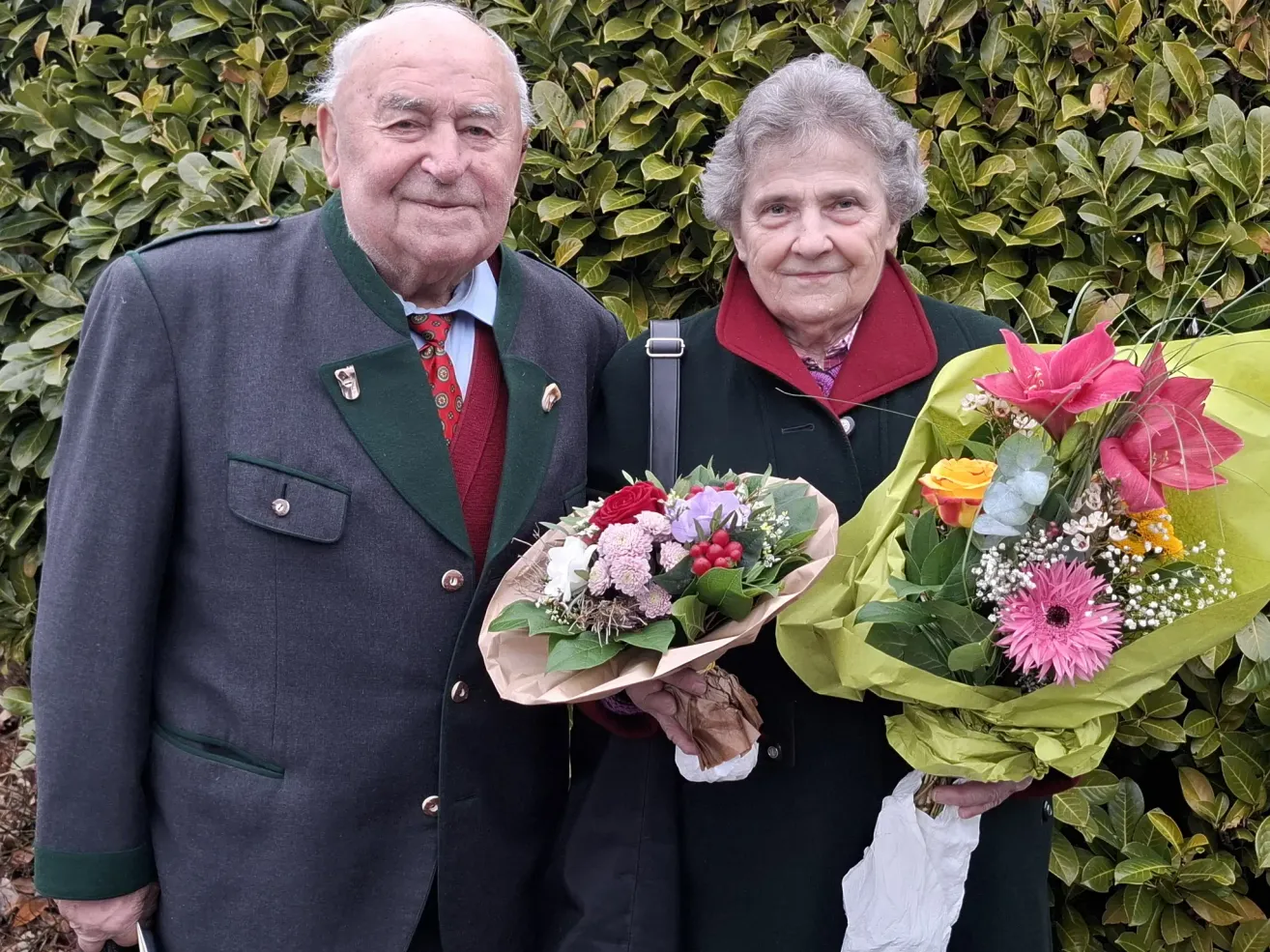 Ein älteres Paar steht lächelnd für ein Foto zusammen und hält Blumensträuße in den Händen. Der Mann trägt eine rote Krawatte und die Frau hat einen roten Kragen. Sie stehen vor einem Busch.