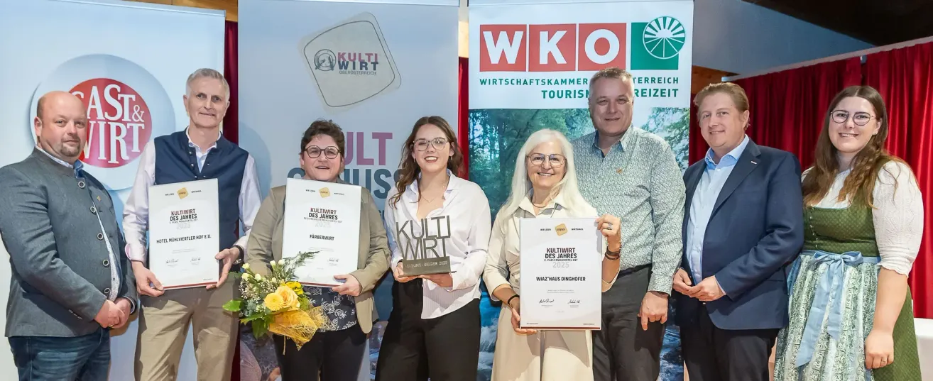 Vier Personen halten Auszeichnungen; eine Frau mit Brille steht lächelnd in der Mitte. Im Hintergrund ist 'WKO' und 'WIRTSCHAFTSKAMMER TOURISM' zu sehen. Ein Mann und zwei Frauen rechts, die Frau links hält einen Blumenstrauß.