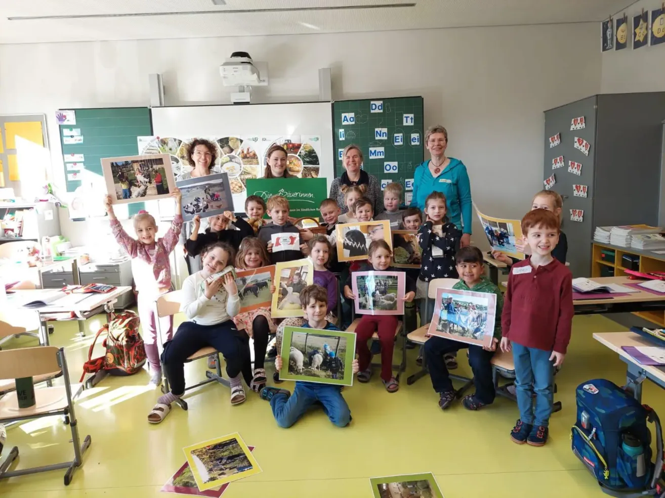 Eine Gruppe von Kindern und Erwachsenen befindet sich in einem Klassenzimmer und hält gerahmte Fotos hoch. Sie sitzen und stehen in verschiedenen Posen. Der Raum ist gut beleuchtet, mit einem gelben Boden.