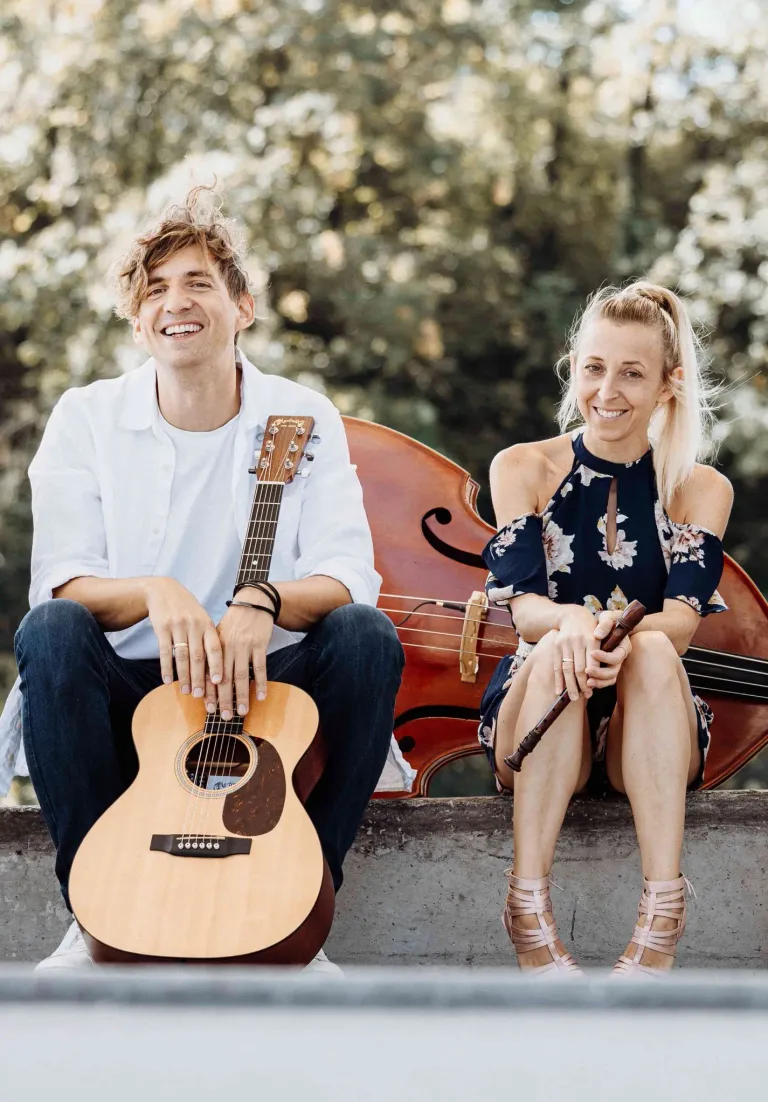 Ein Mann und eine Frau sitzen auf einer Betonbank, lächeln und posieren für ein Foto. Der Mann hält eine Gitarre und die Frau eine Flöte. Sie sind beide lässig gekleidet. Hinter ihnen steht ein großes Cello, und der Hintergrund ist mit Bäumen verschwommen.