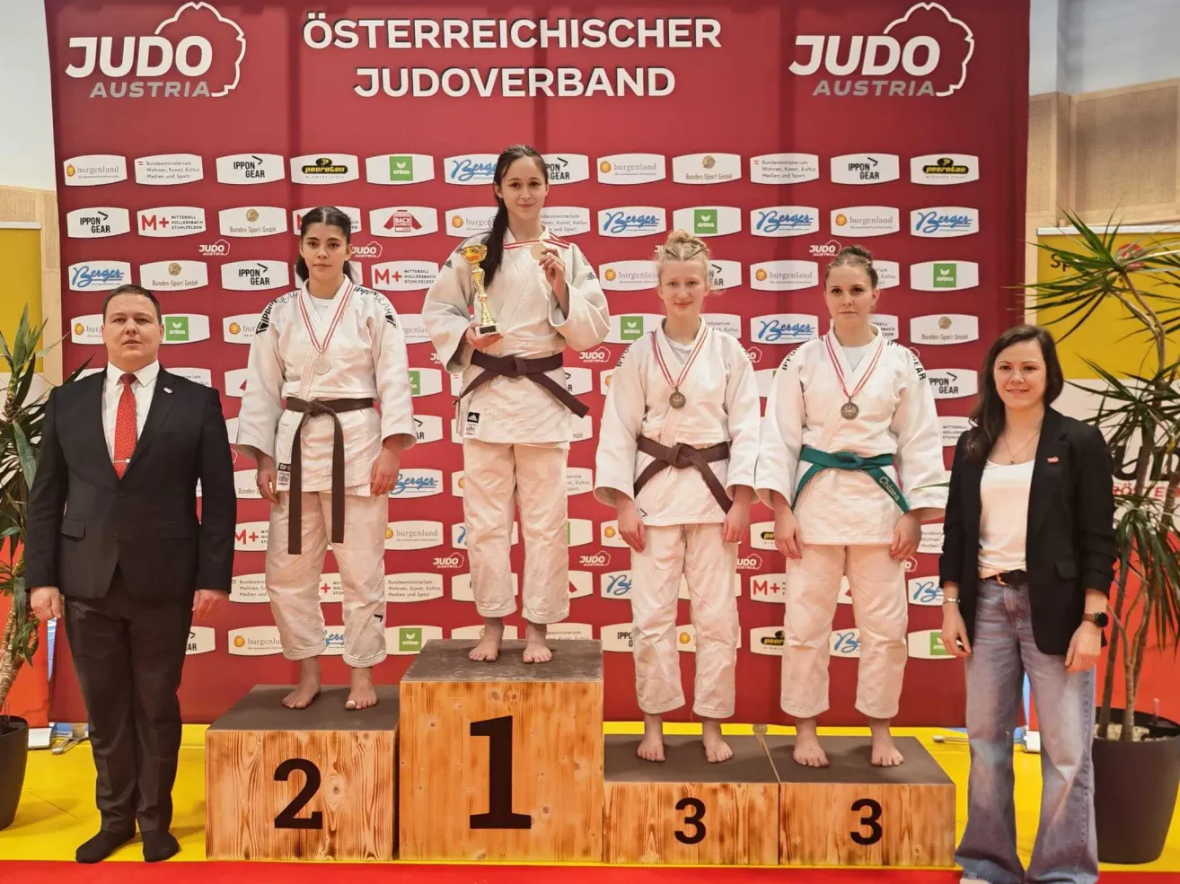 Fünf Frauen in Judo-Uniformen stehen auf einem Podest. Die zentrale Frau hält eine Trophäe. Sie sind von einer roten Wand mit verschiedenen Logos und Sponsoren umgeben. Zwei weitere Personen, eine in formeller Kleidung und die andere in Freizeitkleidung, stehen an den Seiten.