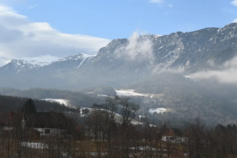 Ein nebeliger Morgenblick auf verschneite Berge mit einem Tal und Häusern darunter, unter einem blauen Himmel mit einigen Wolken.