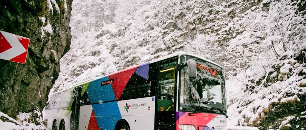 Ein farbenfroher Bus mit der Aufschrift Postbus fährt auf einer verschneiten Bergstraße.
