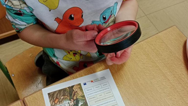 Ein Kind untersucht eine detaillierte Illustration mit einer Lupe, wobei ein Arbeitsblatt mit einem Cartoon-Charakter in der Nähe liegt.