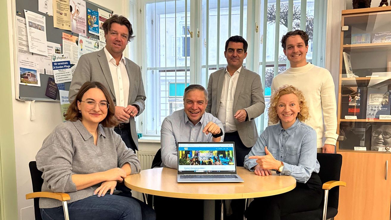 Eine Gruppe von Menschen versammelt sich um einen Laptop, lächelt und posiert für ein Foto in einem Büro. Der Laptop-Bildschirm zeigt eine Website mit Text in einer Fremdsprache an. Hinter ihnen sind Fenster mit weißen Gittern und eine Pinnwand mit Postern zu sehen.