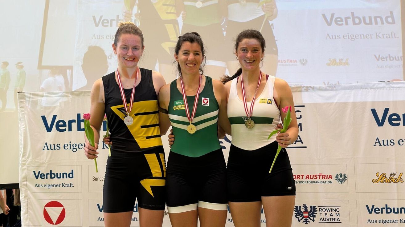 Drei Frauen stehen zusammen und lächeln für ein Foto. Sie tragen Medaillen und halten Blumen, wahrscheinlich zur Feier eines Erfolgs. Ihre Uniformen haben unterschiedliche Farben, und im Hintergrund sind Text und Logos zu sehen.