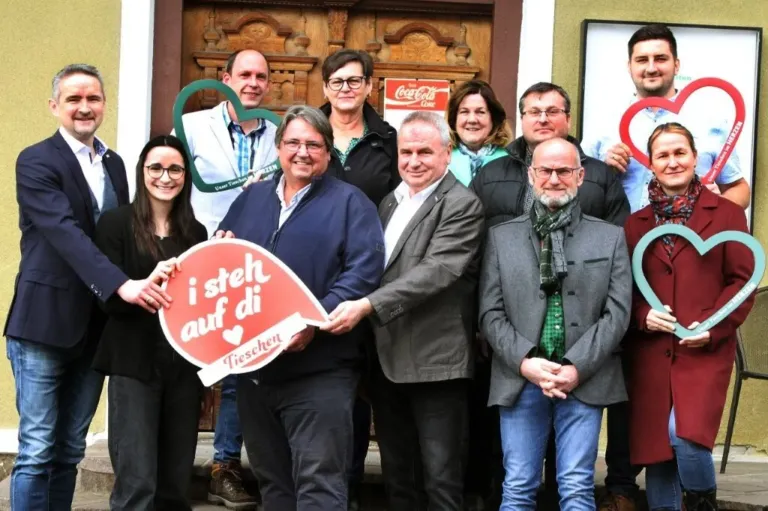 Eine Gruppe von Menschen steht vor einer Holztür. Sie halten ein Herzschild und ein Coca-Cola-Schild. Der Mann in der Mitte hält ein Schild mit der Aufschrift 'i steh auf di'.