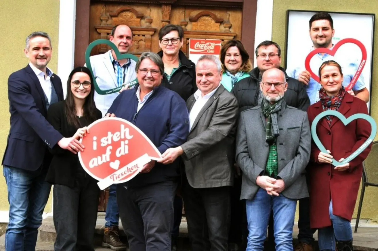 Eine Gruppe von Menschen steht vor einer Holztür. Sie halten ein Herzschild und ein Coca-Cola-Schild. Der Mann in der Mitte hält ein Schild mit der Aufschrift 'i steh auf di'.