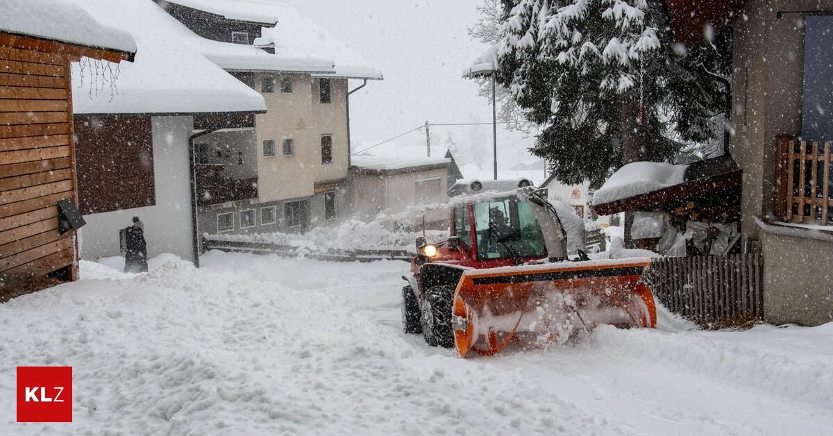 Ein Schneepflug räumt Schnee in einem verschneiten Bereich, mit schneebedeckten Gebäuden und einem schneebedeckten Baum im Hintergrund.
