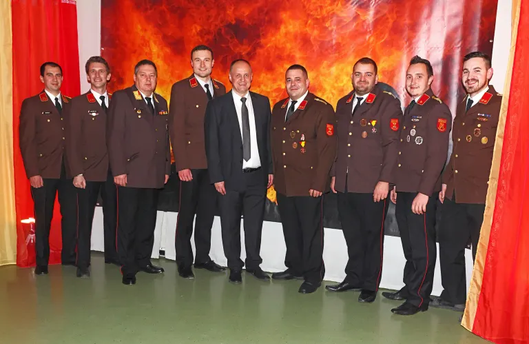 Eine Gruppe von Männern in Militäruniformen steht vor einem Hintergrund aus Feuer, was auf eine formelle Veranstaltung oder Zeremonie hindeutet.