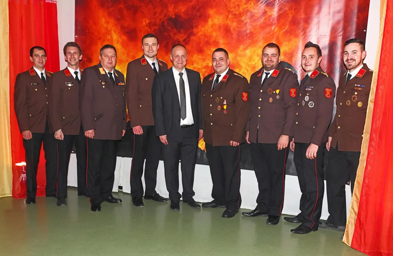 Eine Gruppe von Männern in Militäruniformen steht vor einem Hintergrund aus Feuer, was auf eine formelle Veranstaltung oder Zeremonie hindeutet.