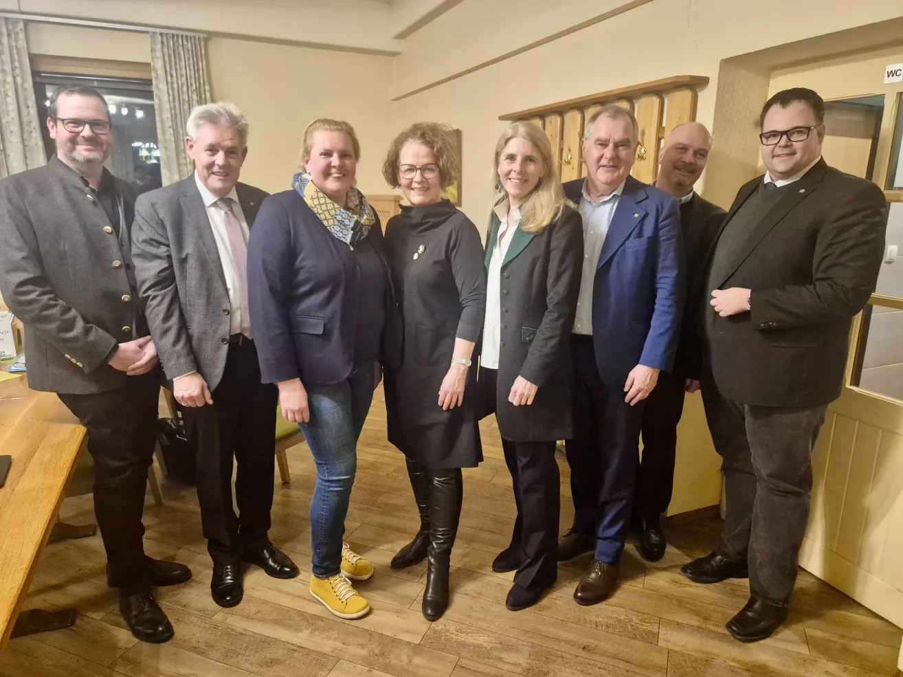 Eine Gruppe von Menschen in formeller Kleidung steht in einem Raum und lächelt für ein Foto. Drei Frauen und drei Männer, von denen zwei eine Brille tragen, stehen in der Mitte. Der Raum hat Holzböden und eine weiße Wand mit einem daran montierten Holzobjekt.