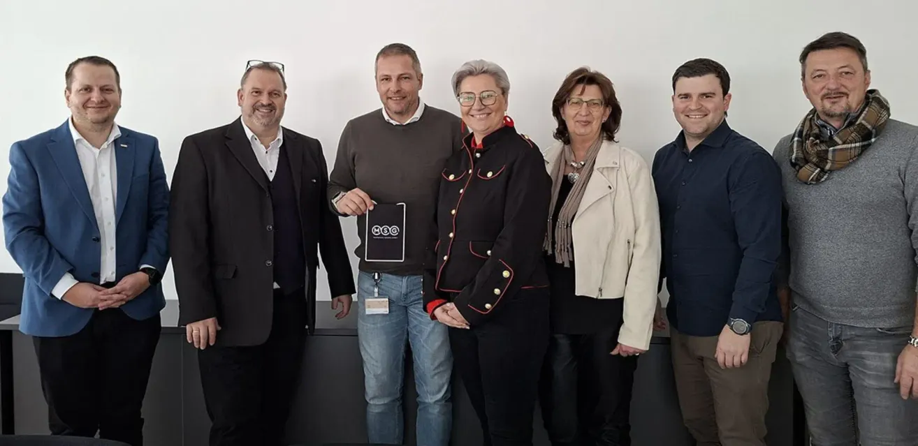 Fünf Erwachsene, drei Männer und zwei Frauen, stehen nebeneinander vor einer weißen Wand. Der Mann in der Mitte hält einen Preis.