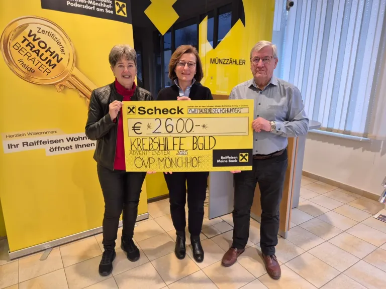 Drei Personen stehen vor einem gelben Hintergrund und halten einen großen gelben Scheck. Die Frau links lächelt, während sie den Scheck hält, und der Mann rechts trägt eine Brille. Dahinter befindet sich ein Fenster mit Jalousien.