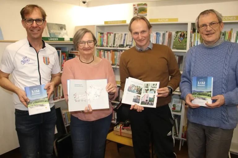 Vier Personen in einer Bibliothek, lächelnd. Drei halten Bücher in den Händen, einer trägt ein weißes Shirt, ein anderer einen braunen Pullover. Im Hintergrund sind mit Büchern gefüllte Regale zu sehen.