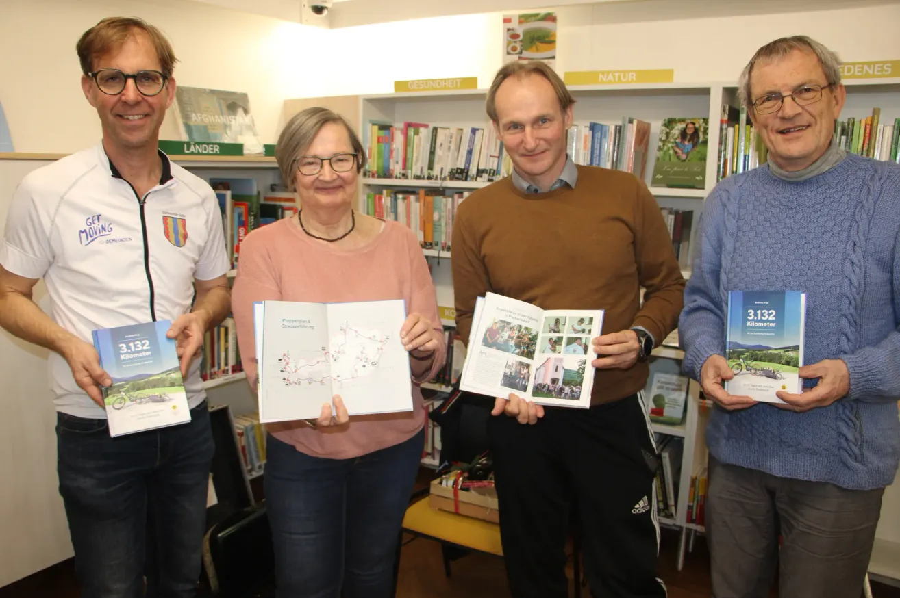 Vier Personen in einer Bibliothek, lächelnd. Drei halten Bücher in den Händen, einer trägt ein weißes Shirt, ein anderer einen braunen Pullover. Im Hintergrund sind mit Büchern gefüllte Regale zu sehen.