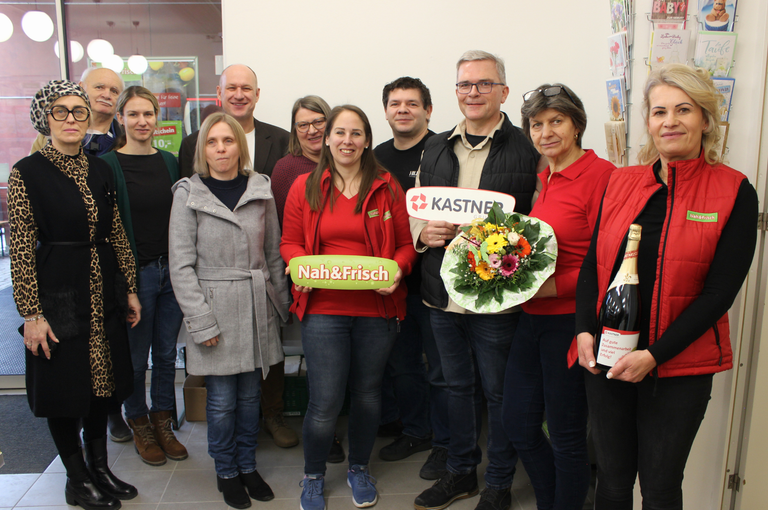 Eine Gruppe von Menschen posiert für ein Foto in einem Büro. Eine Frau in einer roten Jacke hält eine grüne Box mit 'Nah&Frisch' darauf. Hinter ihr hält ein Mann einen Blumenstrauß. Ein anderer Mann hält ein Schild mit 'KASTNER'. Die Leute lächeln.