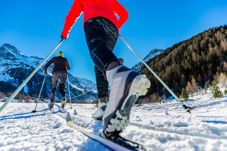 Zwei Skifahrer bewegen sich auf verschneiter Piste, einer in einem roten und der andere in einem schwarzen Jackett, mit Bergen im Hintergrund.