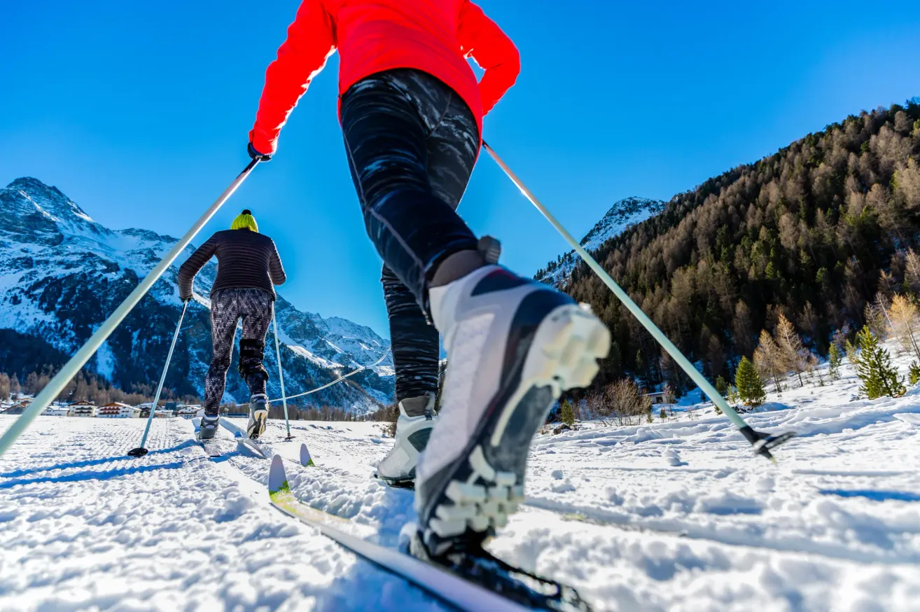 Zwei Skifahrer bewegen sich auf verschneiter Piste, einer in einem roten und der andere in einem schwarzen Jackett, mit Bergen im Hintergrund.