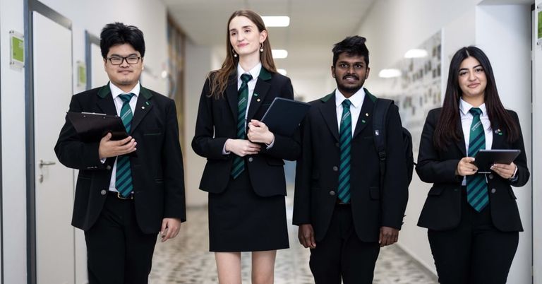 Vier Schüler, zwei Jungen und zwei Mädchen, in formellen Schuluniformen, gehen einen Flur entlang und halten Bücher und Tablets in den Händen.