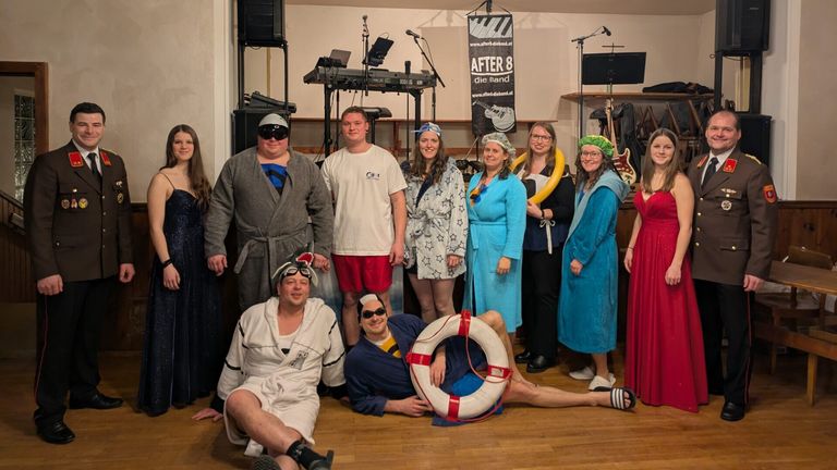 Eine Gruppe von Menschen in Kostümen posiert für ein Foto in einem Raum mit Musikinstrumenten und einem Banner, das 'After 8 die Band' besagt.