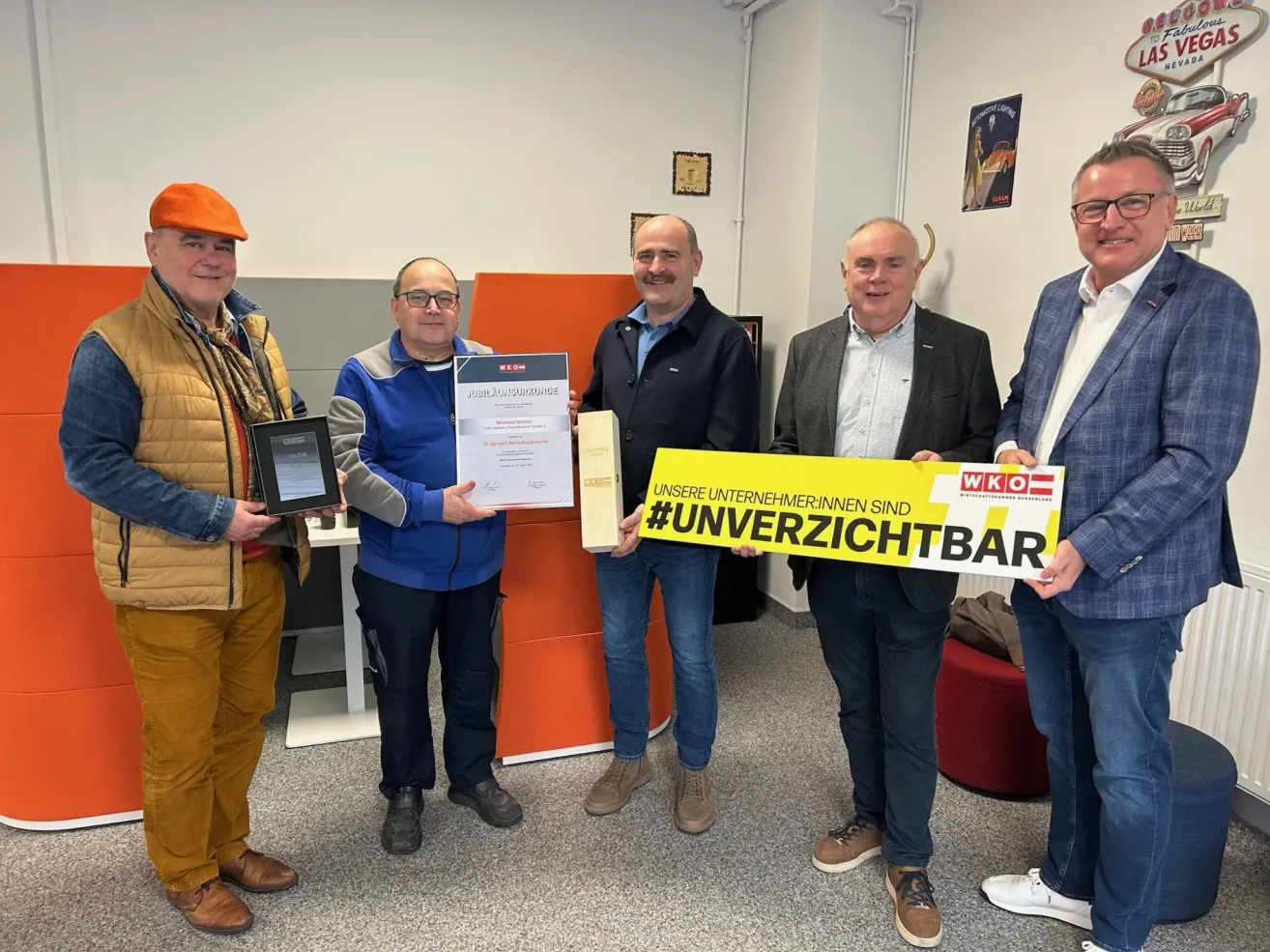 Fünf Männer posieren für ein Foto in einem Büro. Der Mann auf der linken Seite hält einen gerahmten Preis. Der Mann in der Mitte hält ein gelbes Banner. Der Mann auf der rechten Seite hält ein gerahmtes Zertifikat.