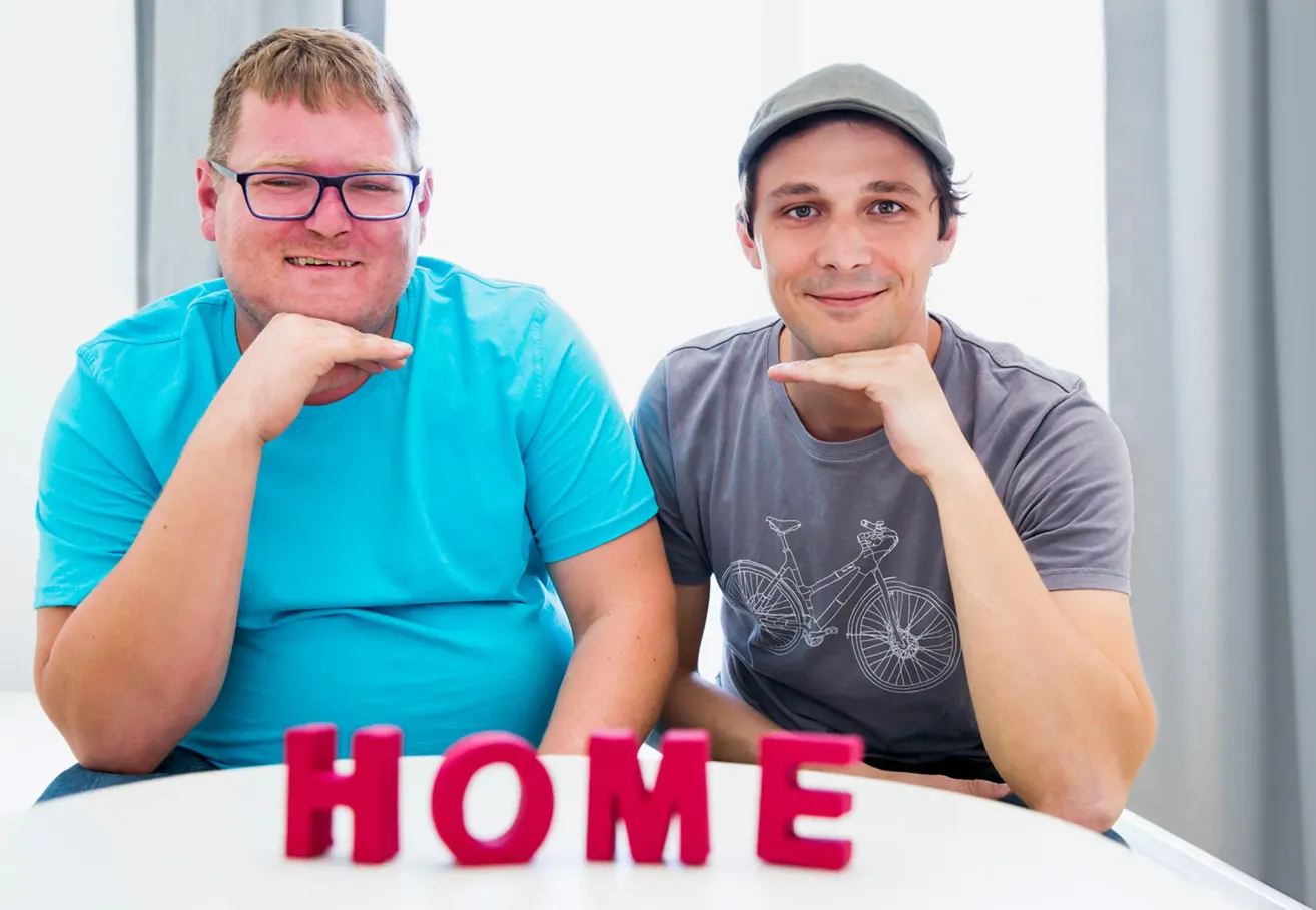 Zwei Männer sitzen nebeneinander, lächeln, einer mit Brille und der andere trägt eine Kappe. Sie posieren mit dem Wort HAUSE auf dem Tisch vor ihnen.