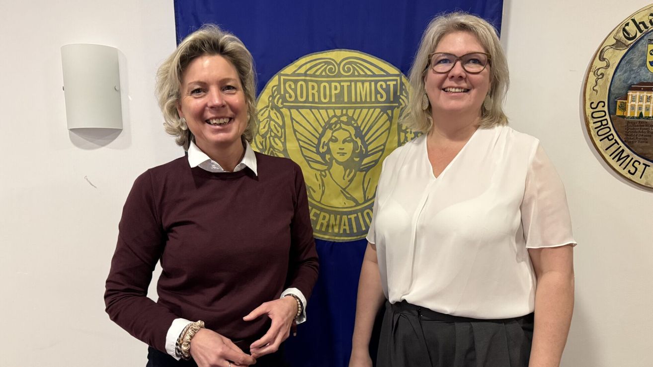 Zwei Frauen stehen vor einer blauen Fahne mit einem gelben Logo, eine trägt ein kastanienbraunes Langarmshirt und die andere ein weißes Blusen. Beide lächeln und schauen in die Kamera.