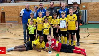 Eine Gruppe junger Fußballspieler mit ihren Trainern posiert für ein Foto in einer Turnhalle. Sie tragen gelbe und schwarze Trikots. Ein Junge in einem roten Trikot hält einen Pokal.