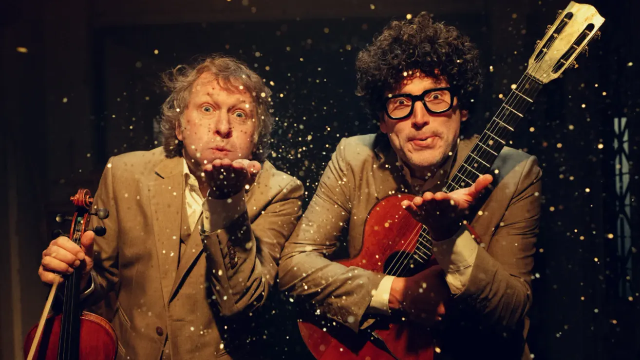 Zwei Männer posieren für ein Foto vor einem glitzernden Hintergrund. Der Mann auf der linken Seite trägt einen Anzug und hat seinen Mund offen. Der Mann auf der rechten Seite trägt eine Brille und hält eine Gitarre.