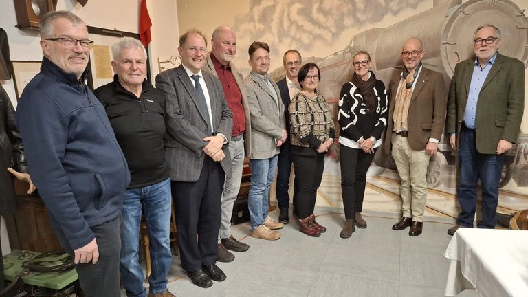 Eine Gruppe Erwachsener steht vor einer Wandmalerei eines Zuges und lächelt für ein Foto. Die Personen sind formell gekleidet, einige tragen Brillen und Schals.