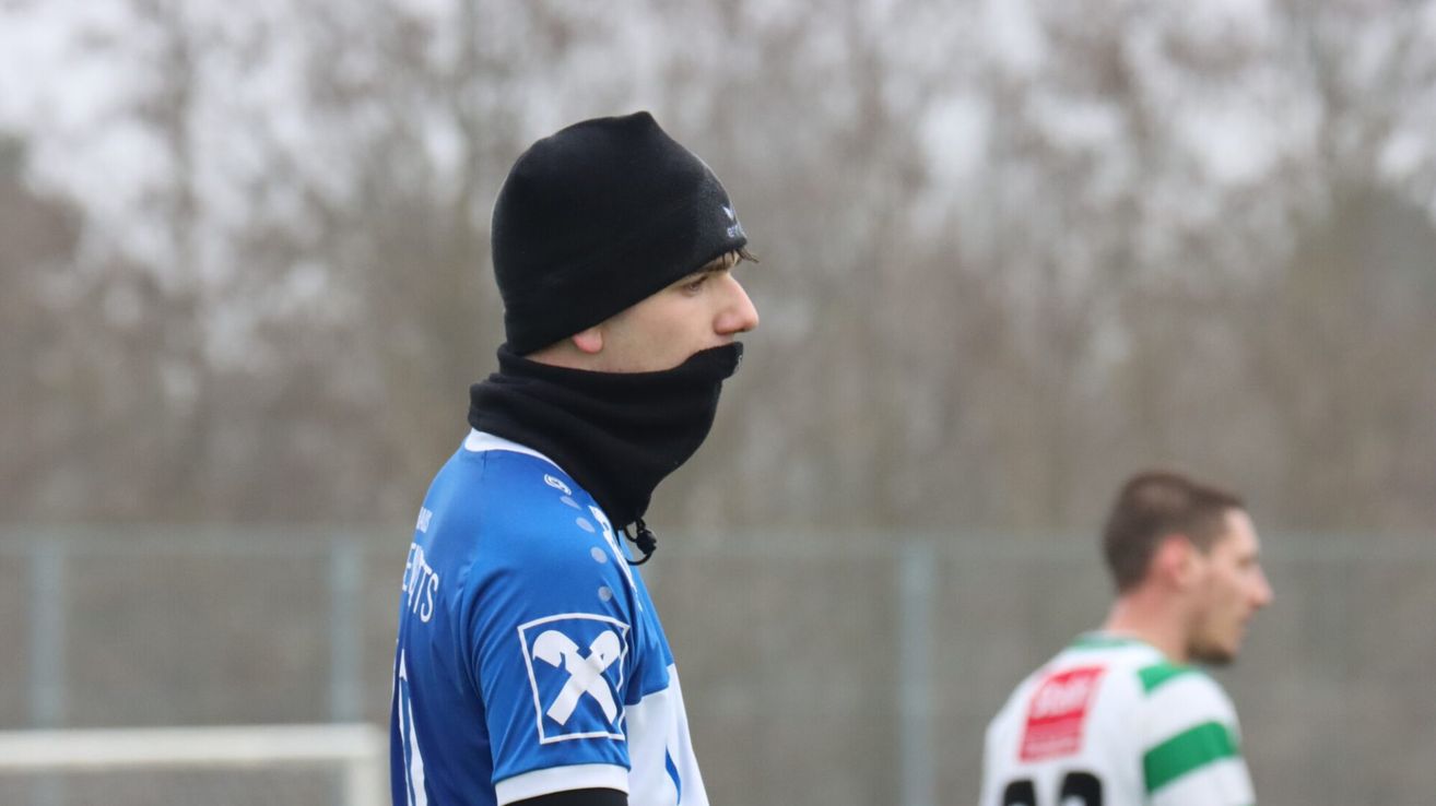 Ein Mann in einem blauen Trikot mit einem weißen X-Logo steht im Freien. Er trägt eine schwarze Mütze und ein Gesichtstuch. Dahinter ist das Trikot einer anderen Person teilweise sichtbar.