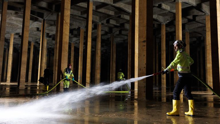 Zwei Arbeiter in Schutzausrüstung reinigen einen überfluteten Bereich mit einem Schlauch in einer Betonstruktur mit hohen Säulen.