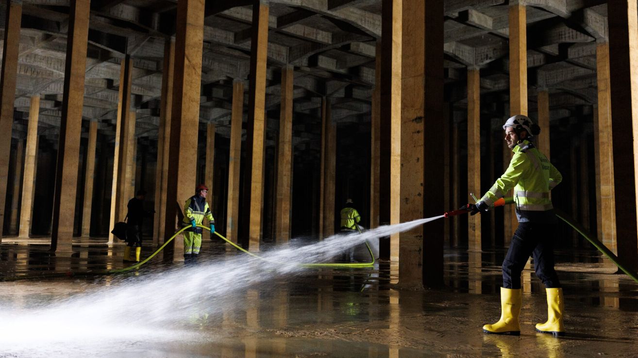 Zwei Arbeiter in Schutzausrüstung reinigen einen überfluteten Bereich mit einem Schlauch in einer Betonstruktur mit hohen Säulen.