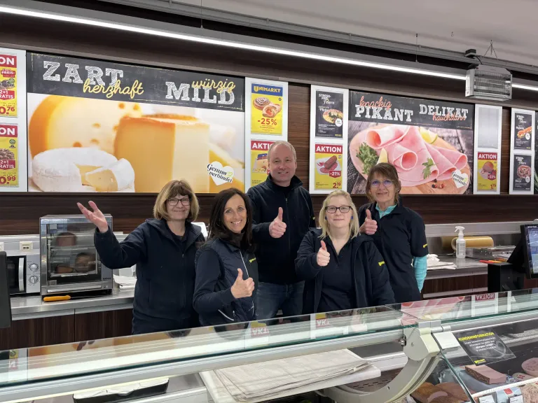 Eine Gruppe von fünf Erwachsenen in schwarzen Jacken steht hinter einer Delikatessentheke in einem Geschäft. Sie alle zeigen Daumen hoch. Dahinter befinden sich Schilder, die Käse, Fleisch und andere Produkte bewerben.