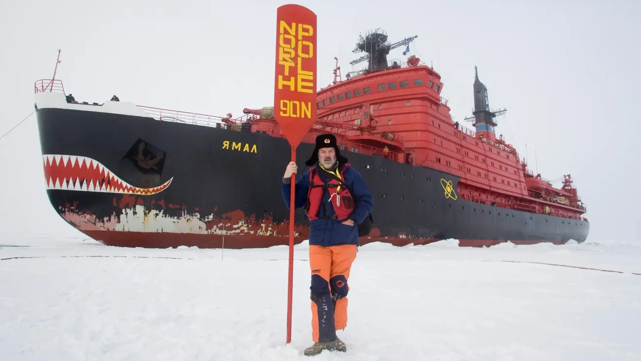 Ein Mann in oranger und blauer Winterkleidung steht im Schnee und hält ein rotes Paddel mit der Aufschrift 'NORDPOL SOHN'. Im Hintergrund steht ein großes rotes Schiff.