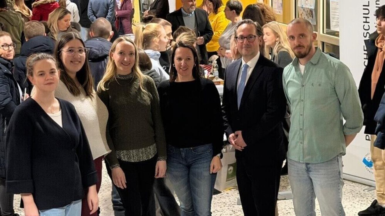 Mehrere Menschen stehen in einem Raum zusammen und lächeln für ein Foto. Sie sind in formeller Kleidung gekleidet. Es gibt einen Mann mit Brille und eine Frau mit einer Halskette.