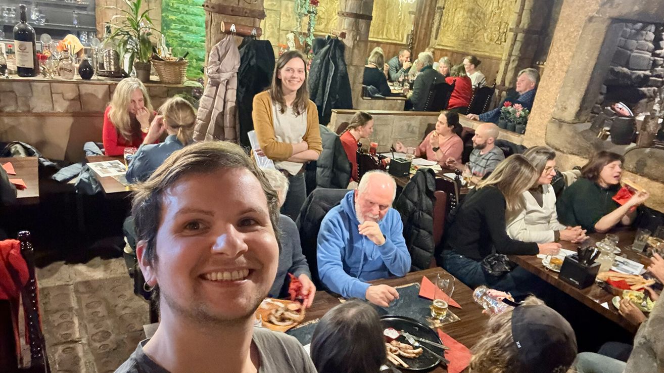 Eine Gruppe von Menschen in einem Restaurant, einige sitzen und andere stehen, mit Essen und Getränken auf den Tischen. Ein lächelnder Mann macht ein Selfie.