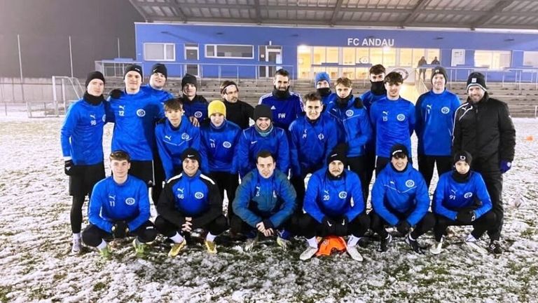 Eine Fußballmannschaft in blauen Trikots posiert für ein Foto vor einem Stadion mit einer blauen Wand und 'FC ANDAU' darauf geschrieben.
