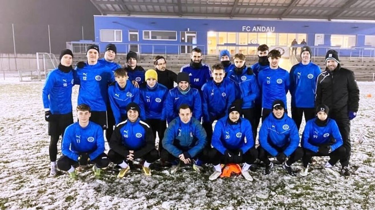 Eine Fußballmannschaft in blauen Trikots posiert für ein Foto vor einem Stadion mit einer blauen Wand und 'FC ANDAU' darauf geschrieben.