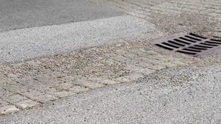 Ein Nahaufnahme einer gepflasterten Straße mit einem Bordstein und einem Abflussdeckel, mit verstreutem Kies und einigen kleinen grünen Grasflecken.
