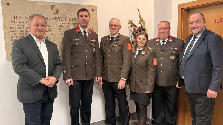 Fünf Personen in Militäruniformen posieren für ein Foto vor einer Gedenkwand mit historischen Daten und einer Statue.